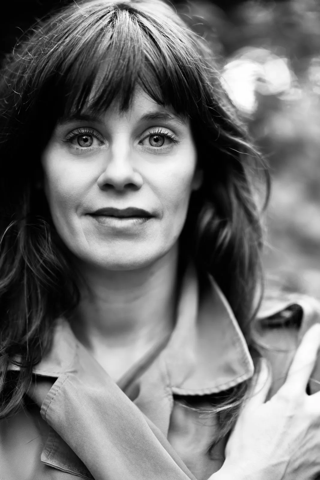 Black-and-white portrait of Tini Lazar with a fringe and loose hair, wearing a trench coat
